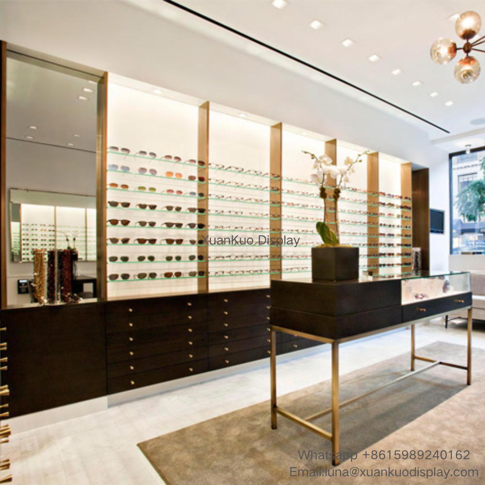 luxury eyewear display furniture set, dark wood multi-drawer base wall cabinet with bronze metal vertical frames, backlit layered glass shelves for optical frames, gold metal leg glass-top central display counter, large mirror panel for high-end optical boutique